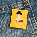 Small rectangular yellow and gold enamel pin with a reimagined gemoetric portrait of Adele Bloch-Bauer I by Gustav Klimt, with gold glitter, pinned on denim fabric.