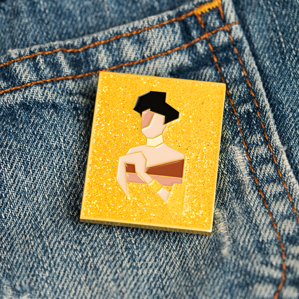 Small rectangular yellow and gold enamel pin with a reimagined gemoetric portrait of Adele Bloch-Bauer I by Gustav Klimt, with gold glitter, pinned on denim fabric.